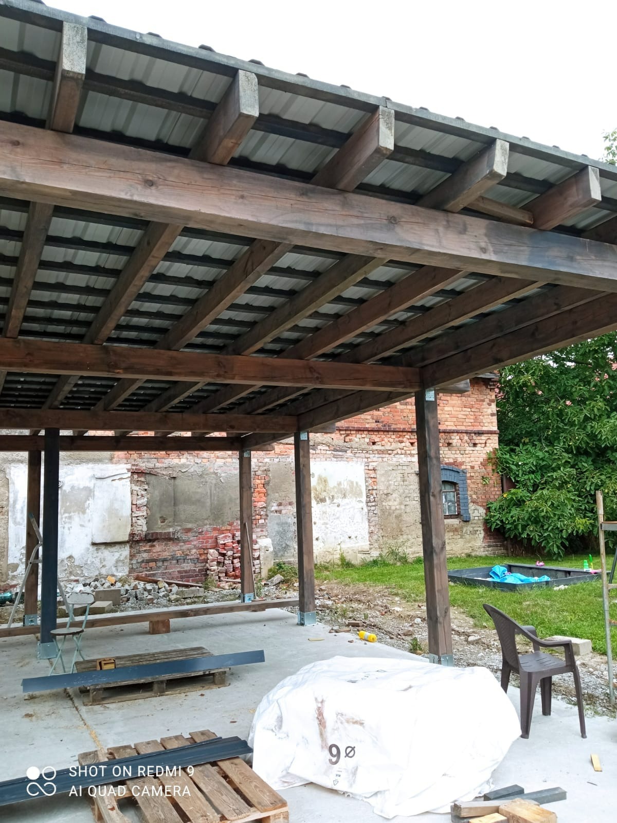 Konstrukcja drewnianej pergoli z metalowym dachem w trakcie budowy, widoczne elementy mocujące, narzędzia i materiały budowlane na betonowej posadzce, w tle ceglany budynek z ubytkami tynku...