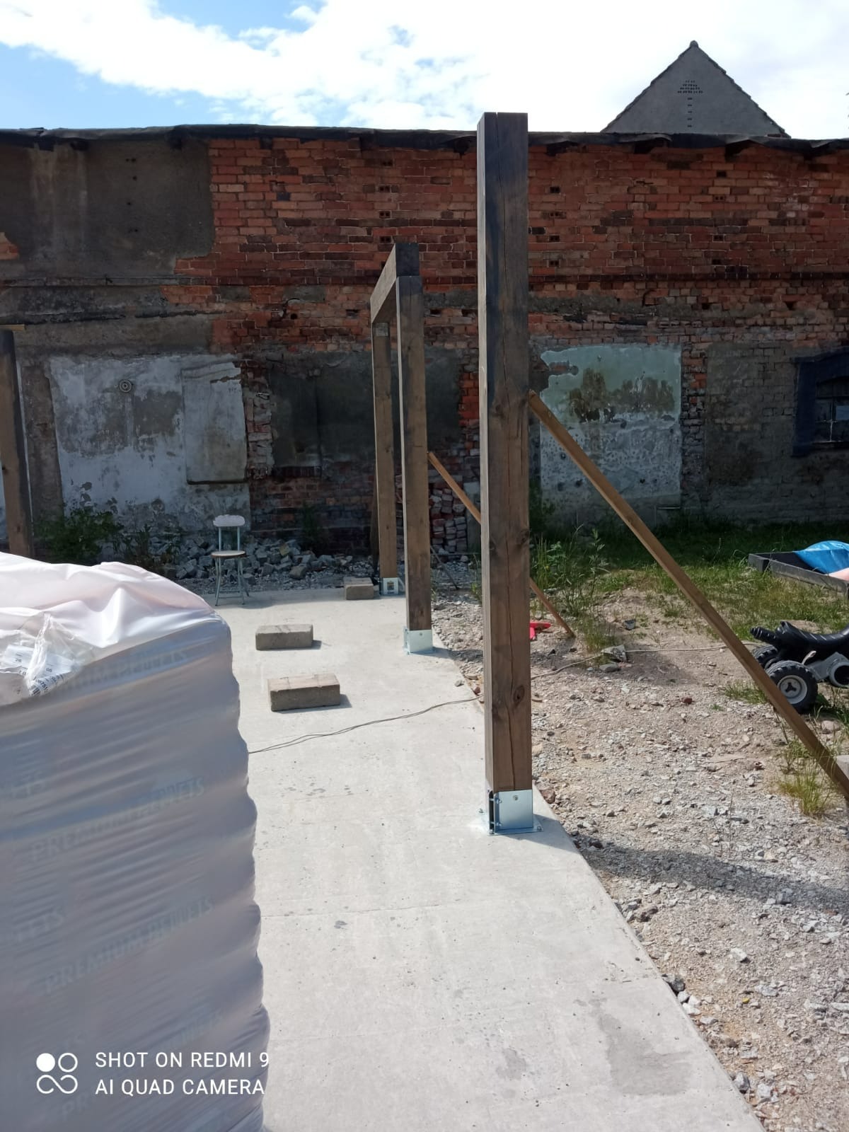 Montaż drewnianej pergoli w trakcie budowy, widoczne słupy z metalowymi mocowaniami, ceglany budynek w tle, przygotowane materiały budowlane obok.
