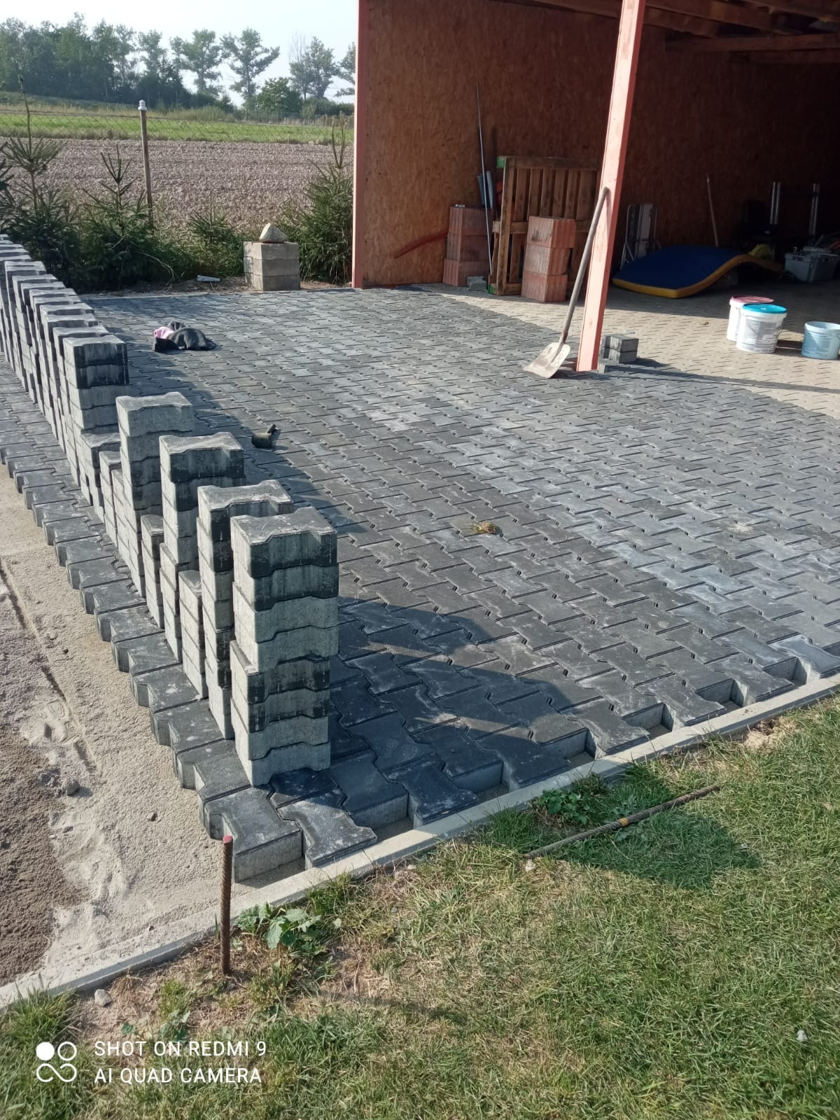 Układanie szarej kostki brukowej na podwórku, widoczne stosy nieułożonych kostek, przygotowane podłoże z piasku, częściowo ukończona nawierzchnia, narzędzia budowlane w tle.