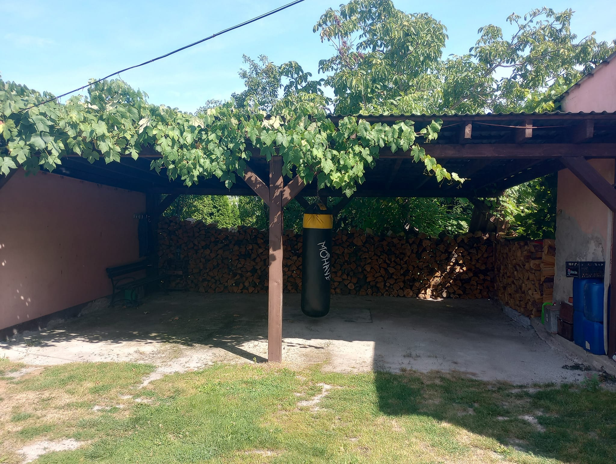 Drewniana wiata z pnącą roślinnością, pod nią stos drewna, worek bokserski i ławka.