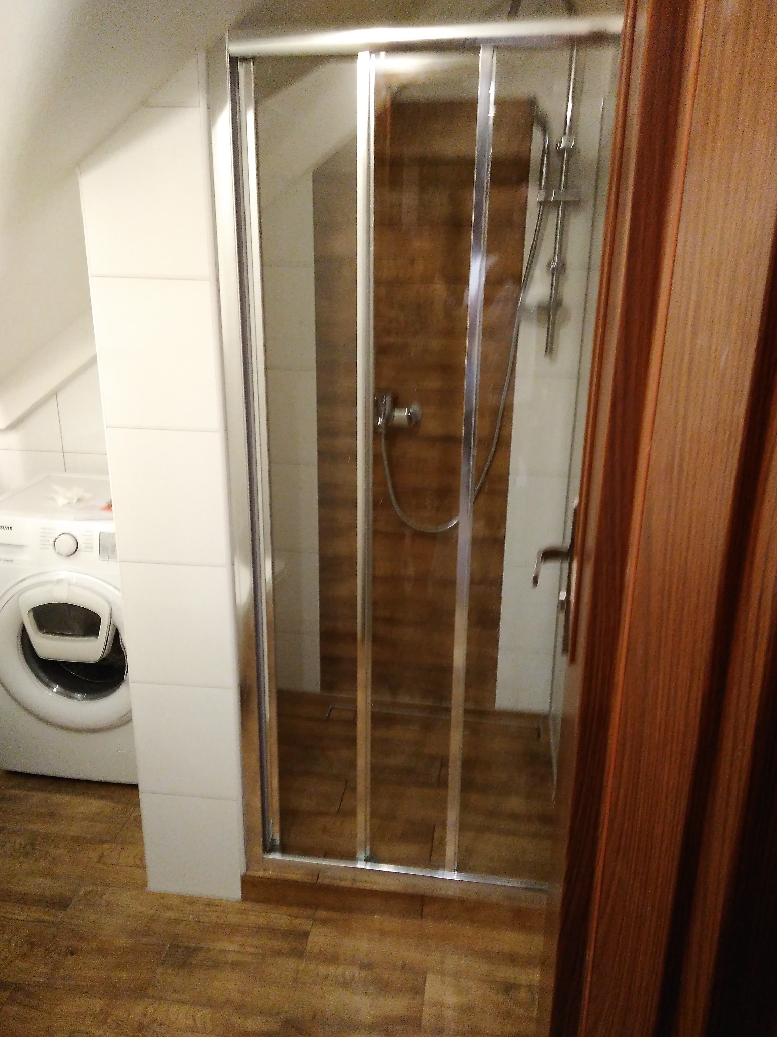 Nowoczesna kabina prysznicowa z przesuwnymi drzwiami, widoczna obok pralka, całość w łazience po remoncie w Ostrowie Wielkopolskim, wykończonej brązowymi płytkami imitującymi drewno.
