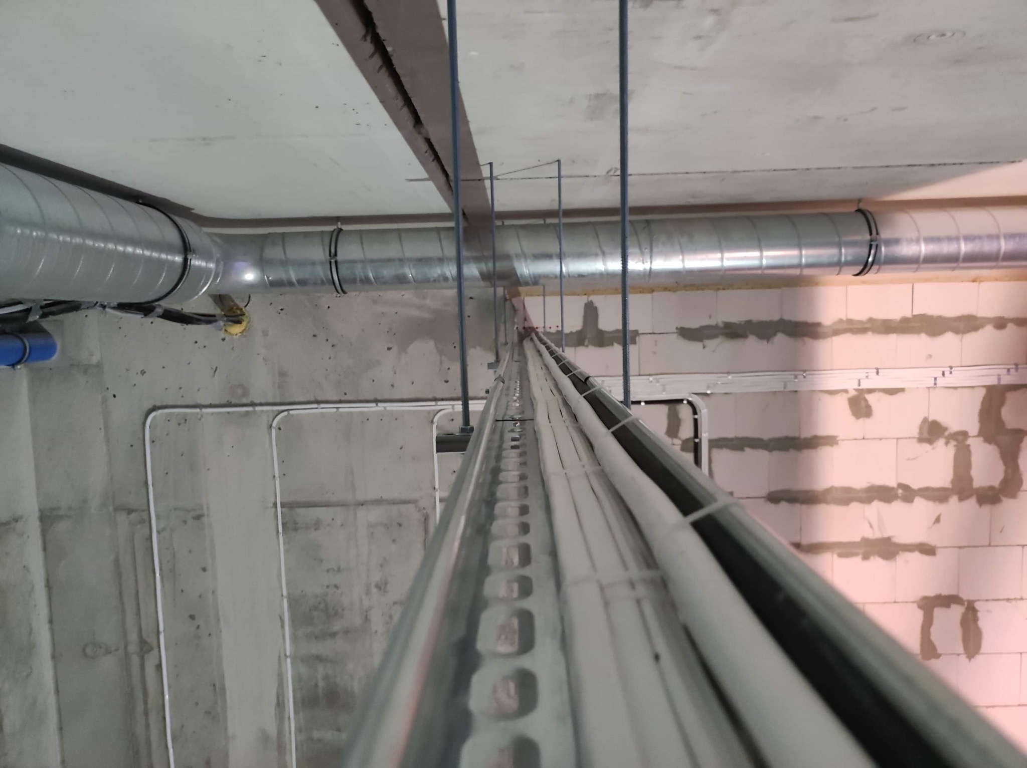 Widok z dołu na koryto kablowe z białymi przewodami, rury wentylacyjne oraz instalacje pod sufitem w surowym stanie budynku.