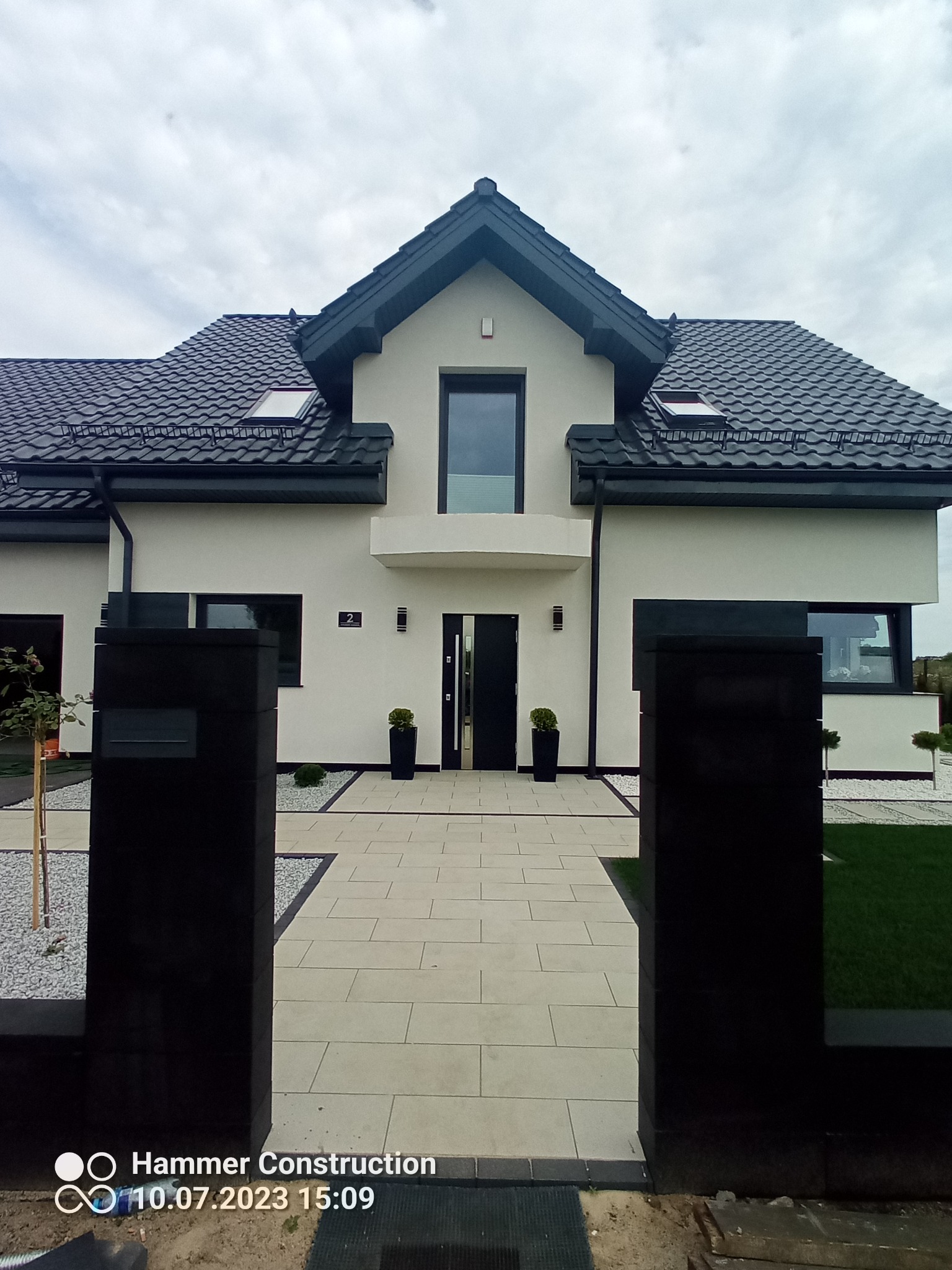 Nowoczesny dom z jasną elewacją, ciemnym dachem i czarnymi akcentami, widok z perspektywy wejścia, z elementami zagospodarowania terenu: trawnik, ścieżka z płyt i ozdobne kamienie.