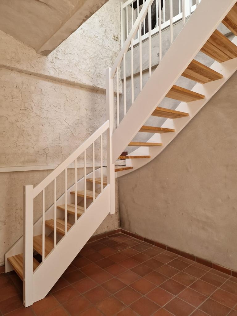 Bieg schodów z drewnianymi stopniami i białą balustradą w surowym wnętrzu z terakotą na podłodze i nierówną fakturą ścian.