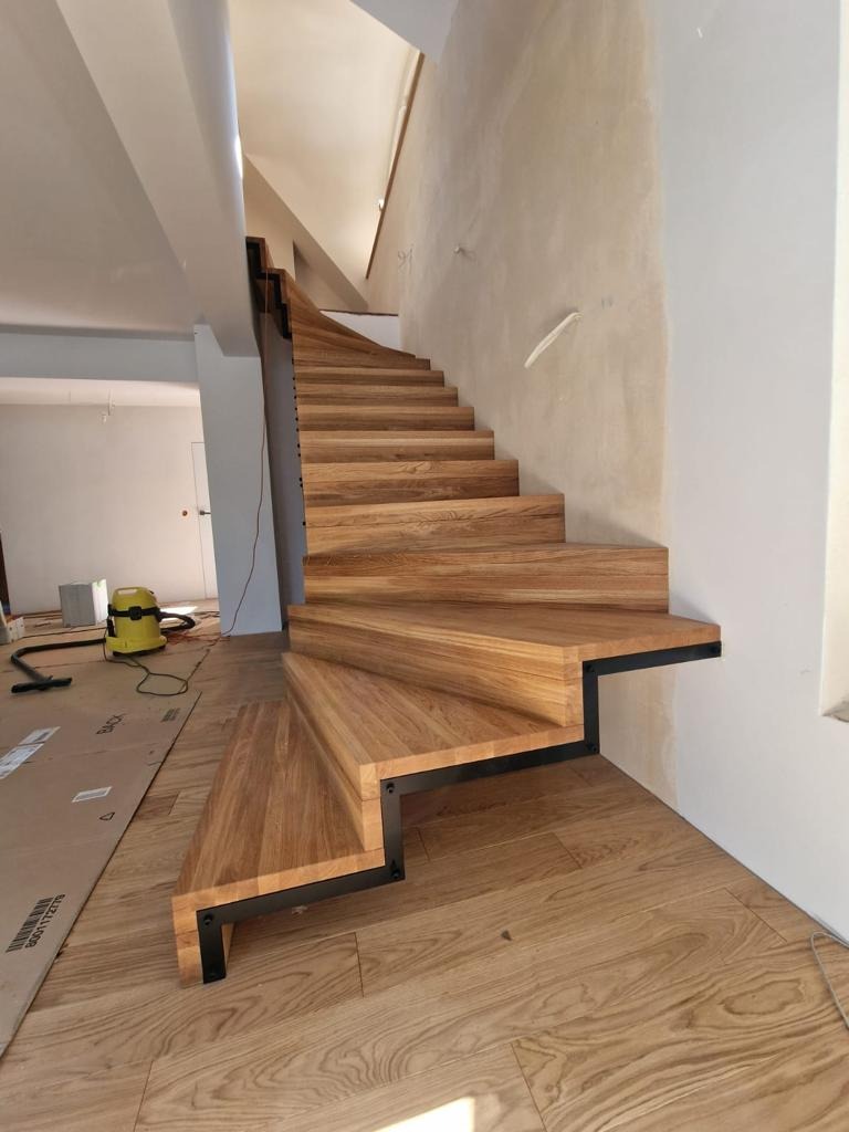 Nowoczesne schody z drewnianymi stopniami i czarną, metalową konstrukcją nośną, widoczne na tle jasnej ściany i podłogi z drewnianych paneli, z odkurzaczem w tle.