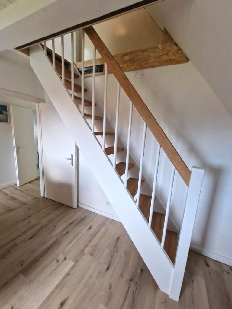 Drewniane schody z białymi balustradami i drewnianą poręczą w jasnym wnętrzu z panelami podłogowymi, widok z dołu.