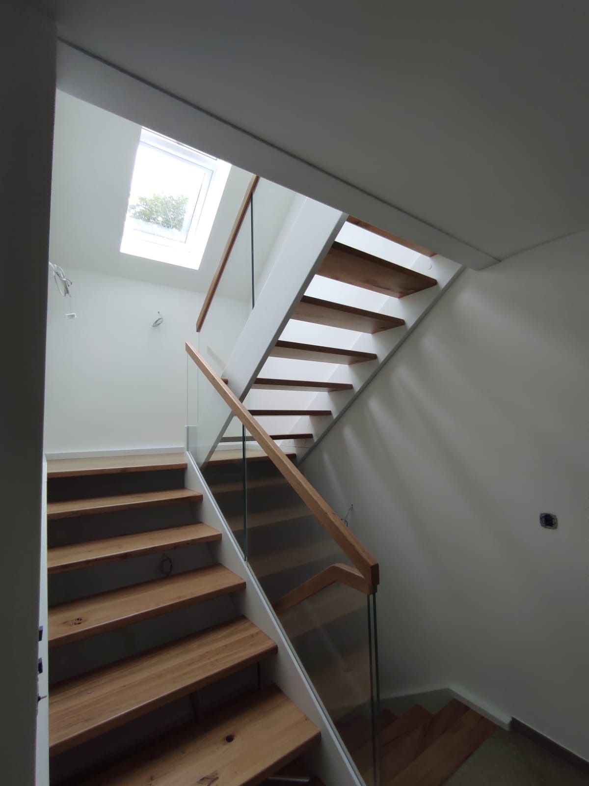 Nowoczesne schody z drewnianymi stopniami i szklaną balustradą, widok z dołu na górę, oświetlone światłem z okna dachowego.