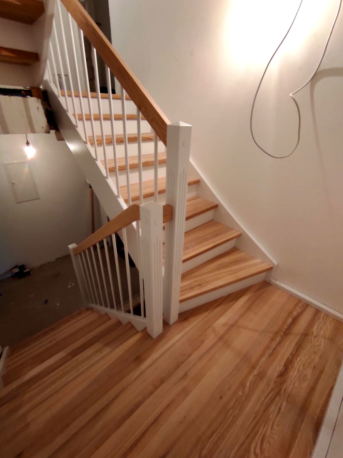 Jasne drewniane schody z białymi balustradami, widok z góry, surowy stan ściany w tle.