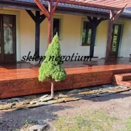 Błyszczący, brązowy taras z drewna, z niskimi schodami i pergolą, otoczony kamieniami i zielenią, przy jasnym budynku.