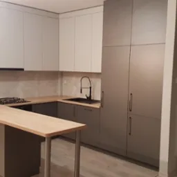 Kuchnia w odcieniach szarości i bieli z drewnianym blatem, widok na zabudowę kuchenną z wyspą i szafami wnękowymi.