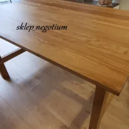 Stół z jasnego drewna z widocznym usłojeniem, stojący na drewnianej podłodze, z delikatnym oświetleniem i znakiem wodnym 'sklep_negotium'.