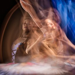 Zuzanna Kulesa - Rozmyte ujęcie baleriny w białym stroju z kolorowymi aplikacjami, uchwycone w dynamicznym ruchu na ciemnym tle, z efektem wielokrotnej ekspozycji i elementami niebieskiej tkaniny.