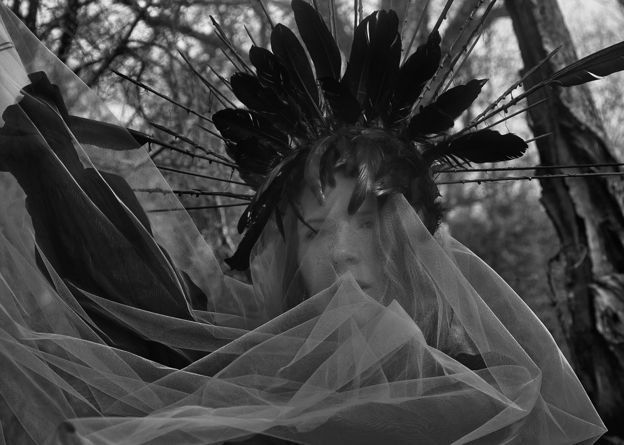 Czarno-białe zdjęcie portretowe osoby z twarzą częściowo zasłoniętą przez tiul, z nakryciem głowy z piór i gałązek na tle drzew.