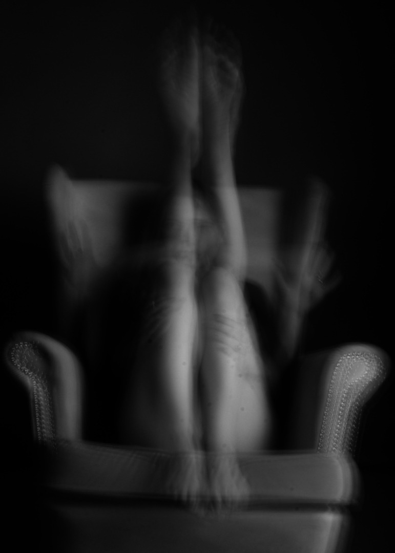 Czarno-białe, rozmyte zdjęcie kobiety siedzącej na fotelu z uniesionymi nogami, twarz zasłonięta dłońmi, artystyczny nieostry efekt.