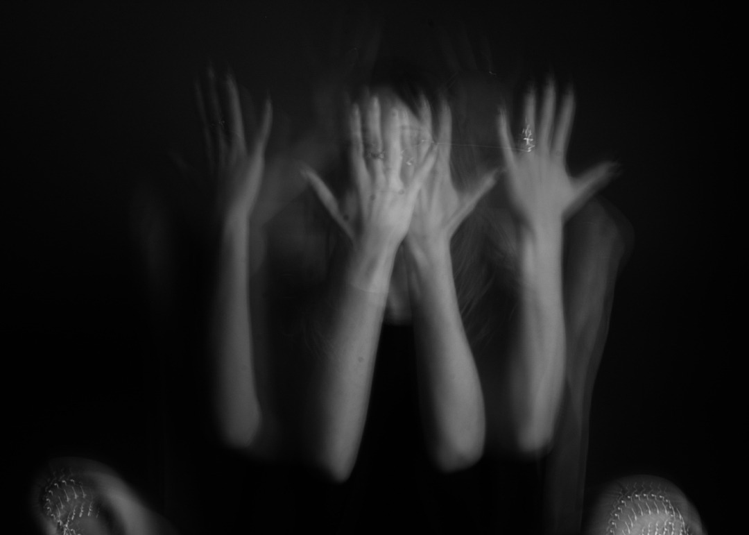 Rozmazane, czarno-białe ujęcie kobiety zakrywającej twarz dłońmi z pierścionkami, efekt ruchu i ciemne tło tworzą intymną atmosferę.
