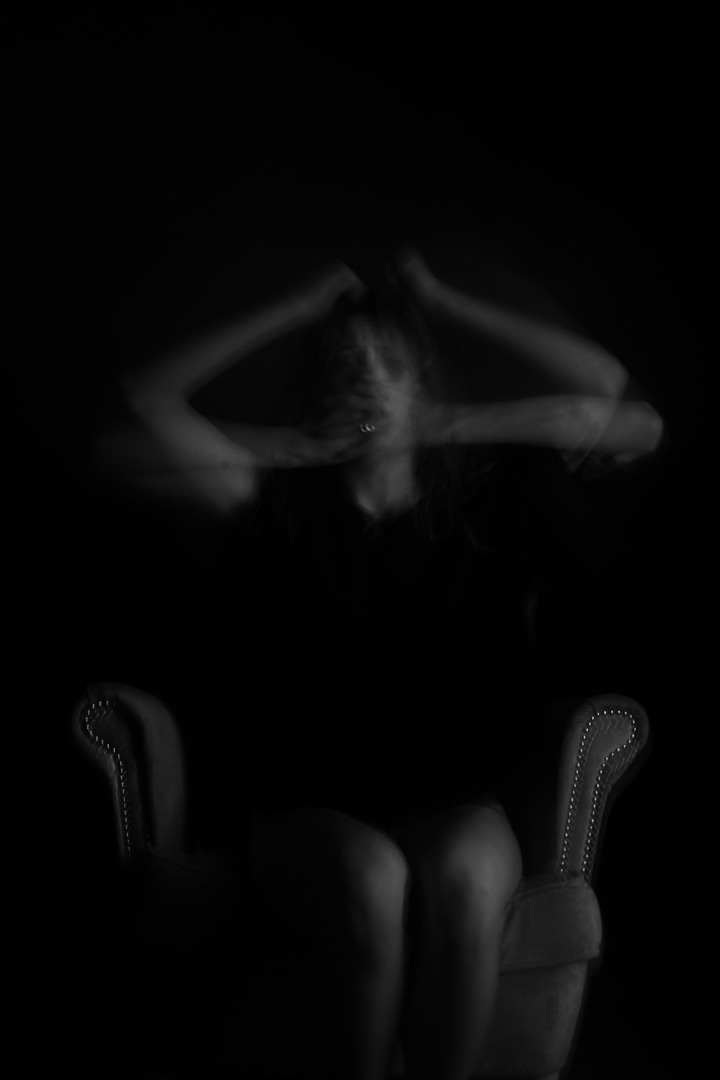 Rozmazane, czarno-białe zdjęcie kobiety siedzącej na fotelu z rękami uniesionymi do głowy, z efektem rozmycia ruchu.