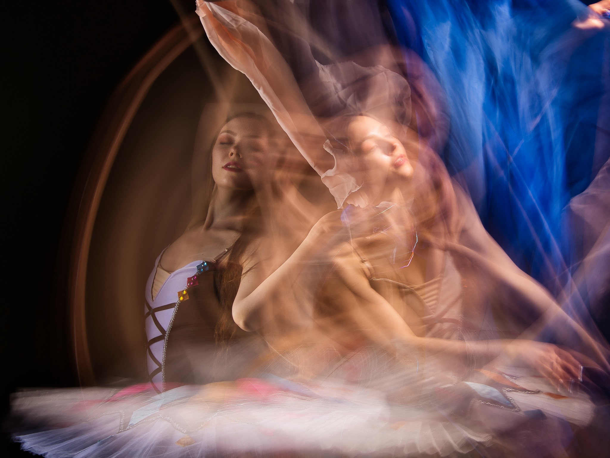 Rozmyte ujęcie baleriny w białym stroju z kolorowymi aplikacjami, uchwycone w dynamicznym ruchu na ciemnym tle, z efektem wielokrotnej ekspozycji i elementami niebieskiej tkaniny.