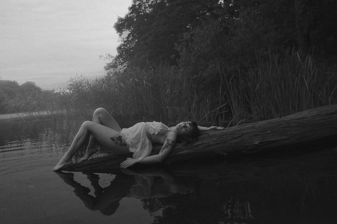 Czarno-biała sesja: Kobieta w koronkowej bieliźnie leży na powalonym pniu drzewa nad wodą, z widocznym tatuażem na udzie i ramieniu.