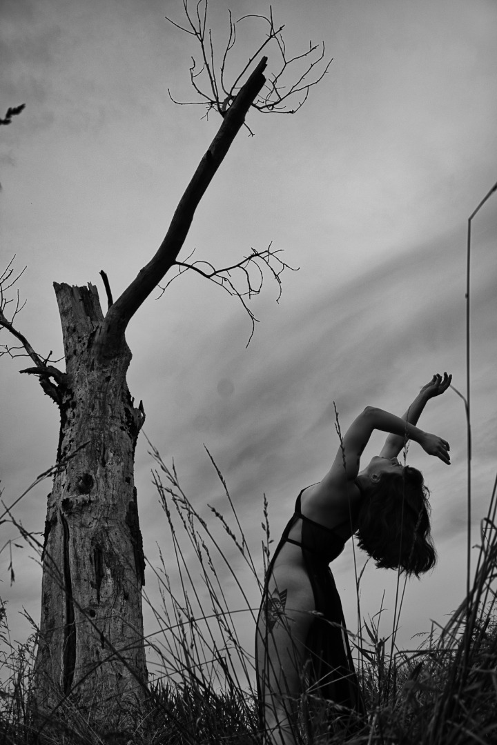 Czarno-białe zdjęcie kobiety w sukience, wyginającej się do tyłu na łące z wysoką trawą, obok uschniętego drzewa z pojedynczą gałęzią, pod pochmurnym niebem.