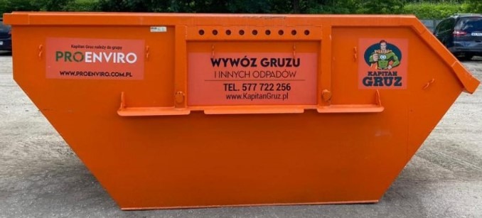 Pomarańczowy kontener na odpady z naklejkami firmowymi ProEnviro i Kapitan Gruz, widoczny na tle zieleni i zaparkowanych aut.
