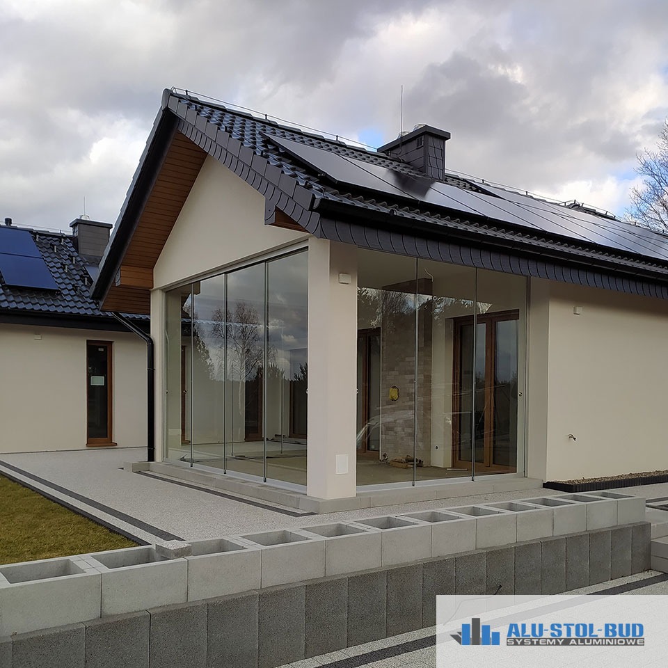 Nowoczesny dom z przeszklonym tarasem i panelami słonecznymi na dachu, widok z zewnątrz na szarą kostkę brukową i elementy betonowe.