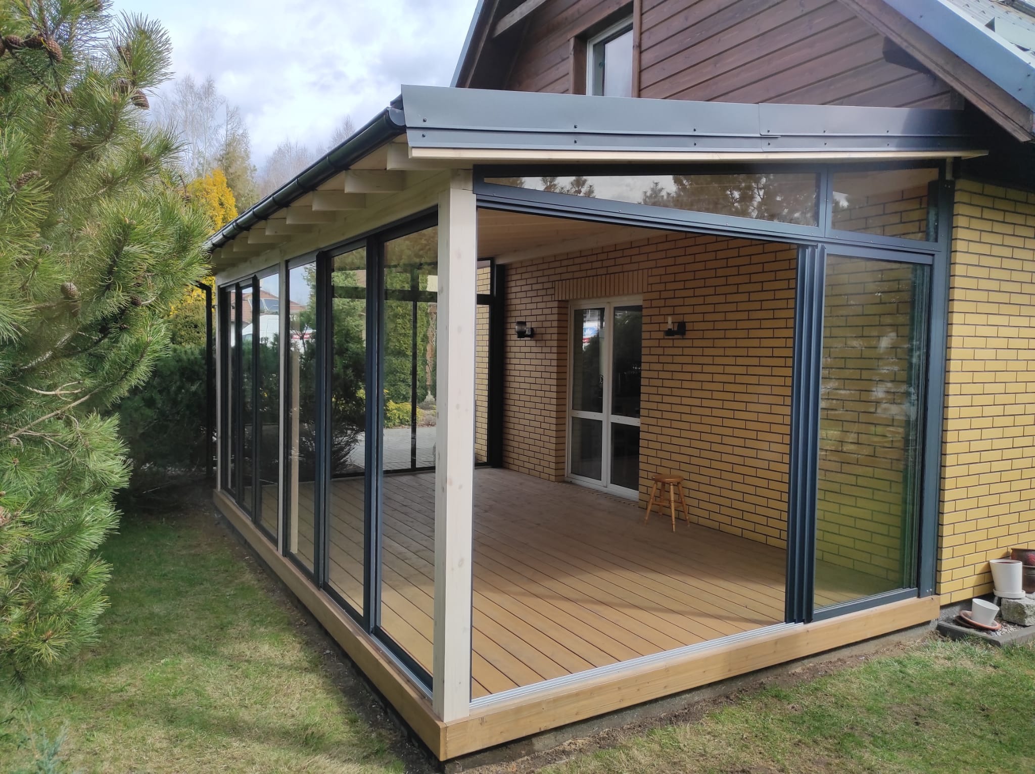 Zadaszony taras z przesuwnymi szklanymi ścianami i kompozytową podłogą, przylegający do ceglanego domu z drewnianą elewacją, widoczny mały drewniany stołek wewnątrz.
