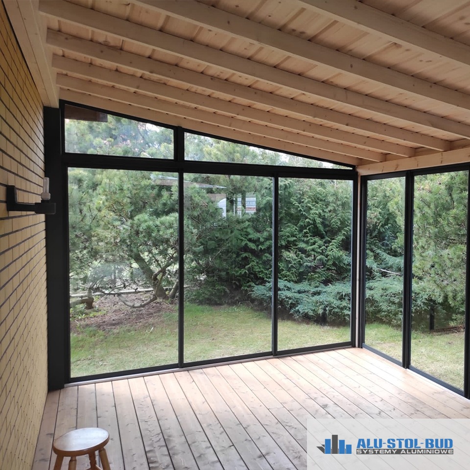 Zadaszony taras z drewnianą podłogą i przeszklonymi ścianami w czarnych ramach, widok na ogród z bujną zielenią, na podłodze mały drewniany taboret, logo firmy w prawym dolnym rogu.