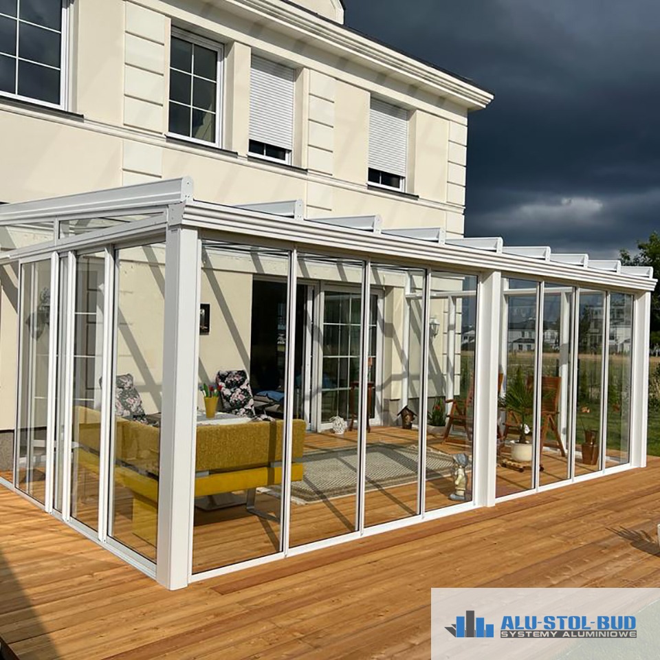 Przeszklona konstrukcja z białymi ramami na drewnianym tarasie, widok na wnętrze z kanapą i dekoracjami, w tle dom z roletami i ciemne chmury.