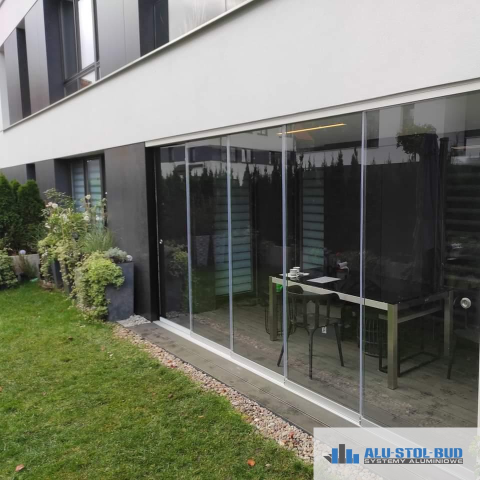 Przesuwne szklane panele tworzące zabudowę tarasu przy nowoczesnym domu z zielonym trawnikiem i roślinami w donicach.