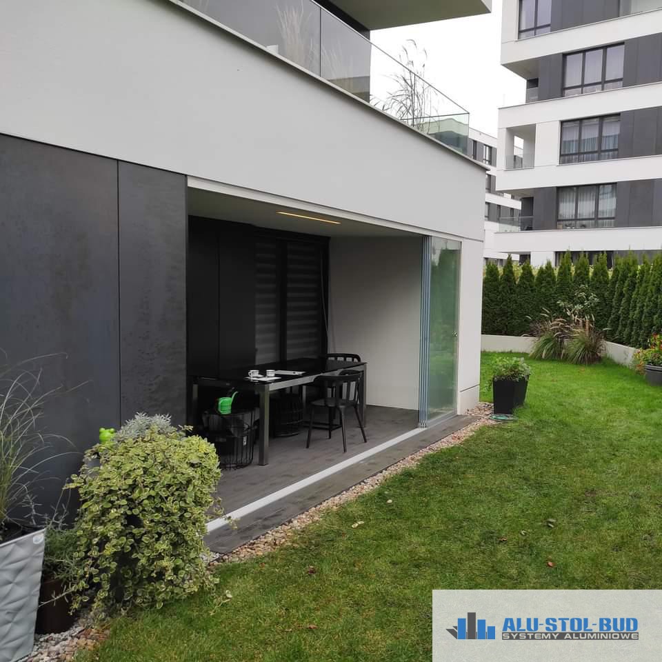 Nowoczesny, zadaszony taras z przesuwnymi szklanymi drzwiami, wyposażony w stół i krzesła, widok z ogrodu na tle budynku mieszkalnego.