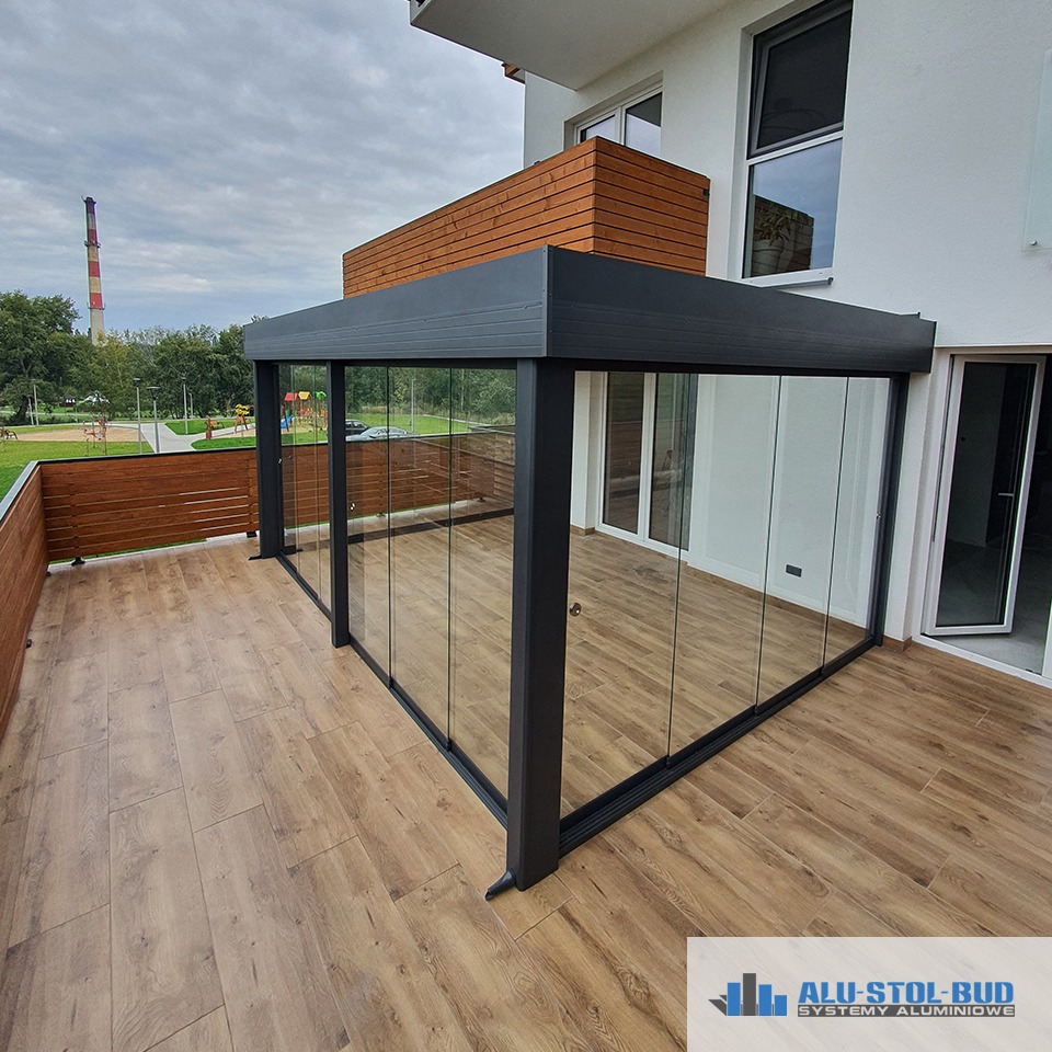 Zadaszony taras z przesuwnymi szklanymi ścianami i podłogą z desek kompozytowych, widok na park i komin w tle. Na dole widoczne logo firmy.