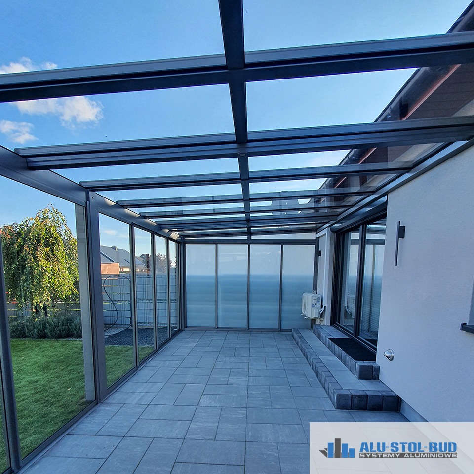 Zadaszony taras z metalową konstrukcją i szklanymi ścianami, widok z zewnątrz na ogród, szara kostka brukowa, logo firmy w rogu.