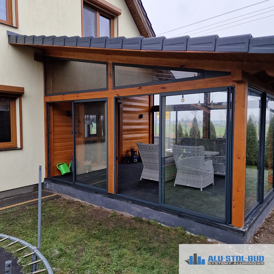 Zadaszony taras z drewnianym wykończeniem, szklanymi ścianami i przesuwnymi drzwiami, widok z zewnątrz na meble ogrodowe w środku i zielony trawnik na pierwszym planie.