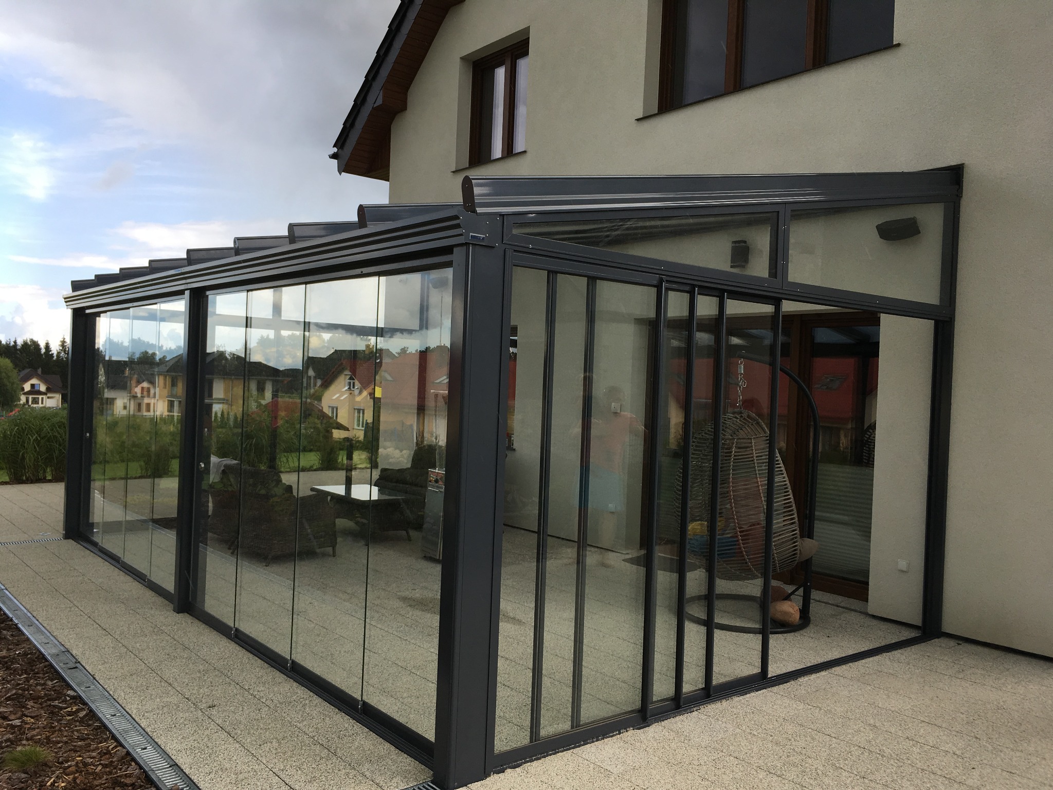 Zadaszony taras z przesuwnymi szklanymi ścianami i metalową konstrukcją, widok na ogród i domy w tle. Wewnątrz tarasu rattanowy fotel wiszący i meble wypoczynkowe.