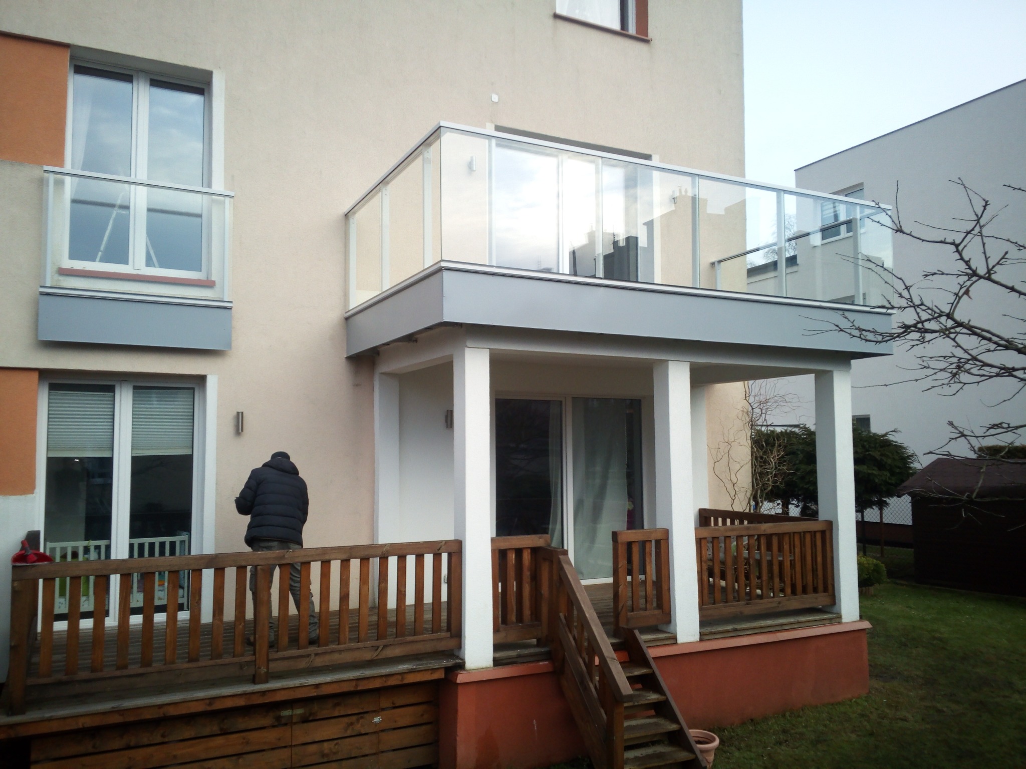 Dwupoziomowy taras przy budynku mieszkalnym z drewnianą balustradą na dolnym poziomie i szklaną na górnym, osoba na tarasie dolnym.