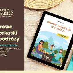 Reklama e-booka o zdrowych przekąskach w podróży, wyświetlana na tablecie leżącym na piasku obok słomkowego kapelusza i klapek.