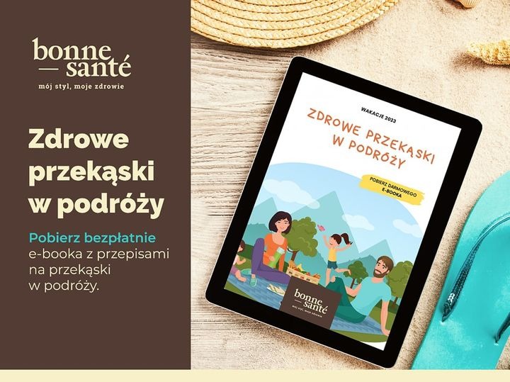 Reklama e-booka o zdrowych przekąskach w podróży, wyświetlana na tablecie leżącym na piasku obok słomkowego kapelusza i klapek.