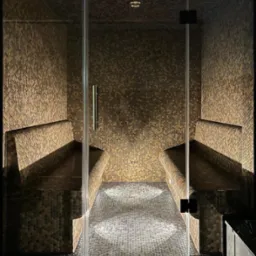 Wnętrze kabiny z mozaiką heksagonalną w odcieniach brązu i szarości, z dwoma ławami po bokach i szklanymi drzwiami, oświetlone punktowo