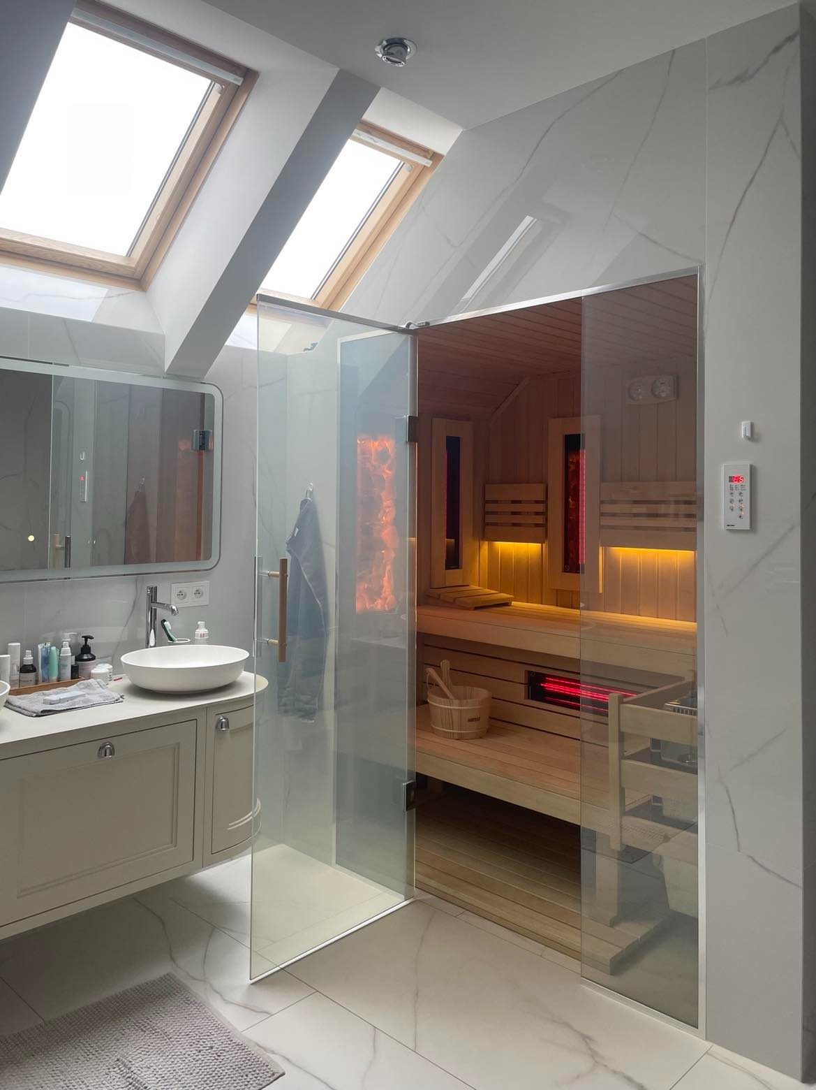 Nowoczesna łazienka z przeszkloną kabiną sauny, widoczne drewniane ławki, oświetlenie LED i piec infrared, całość doświetlona przez okna dachowe.