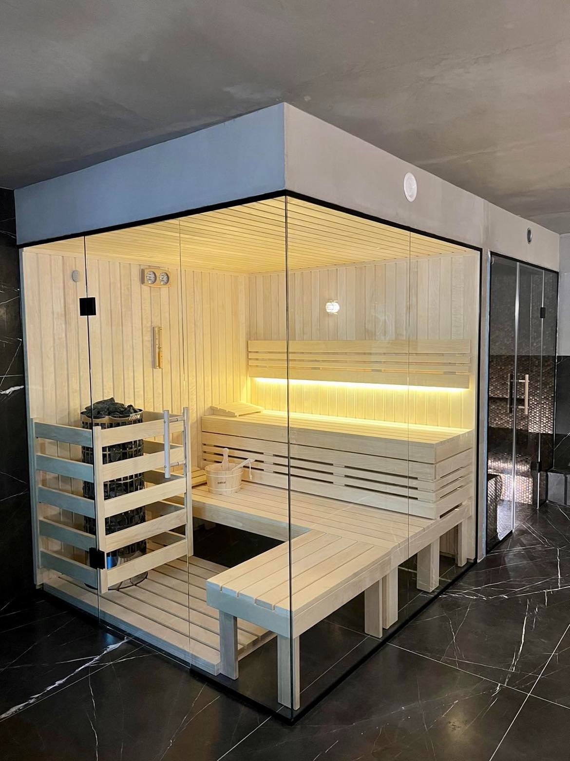 Nowoczesna kabina z jasnego drewna z przeszklonymi ścianami, podświetlone ławy, piec z kamieniami i czarne marmurowe płytki na podłodze.
