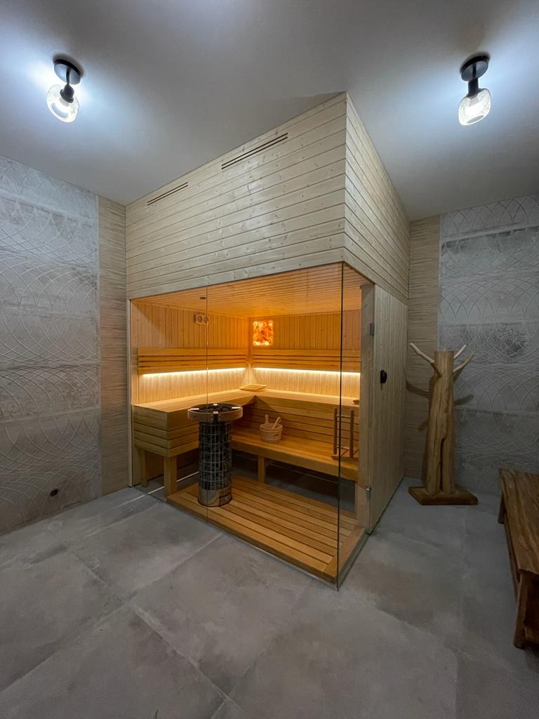 Nowoczesna kabina z drewna z przeszklonymi drzwiami, podświetlane ławki, piec w metalowej obudowie i dekoracja imitująca drzewo w pomieszczeniu wyłożonym płytkami.