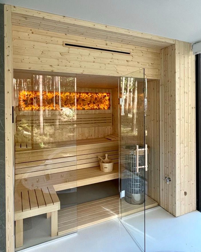 Nowoczesna, jasna kabina z drewna z przeszklonymi drzwiami, podświetlanym panelem z motywem lasu i metalowym piecem wewnątrz, widoczny drewniany ceber.