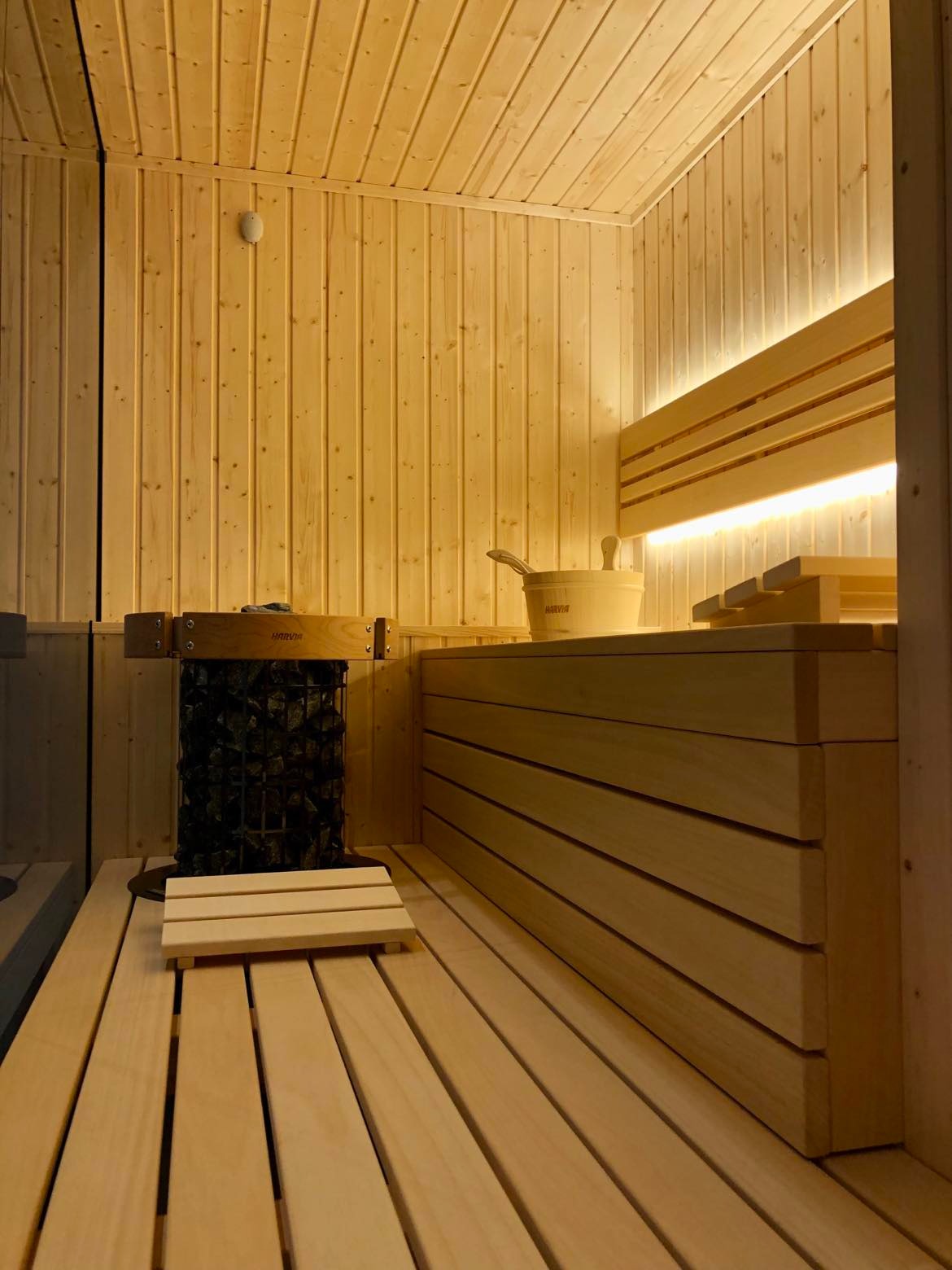 Wnętrze drewnianej kabiny z piecem na kamienie, drewnianym wiadrem z chochlą i podświetlanymi ławkami.