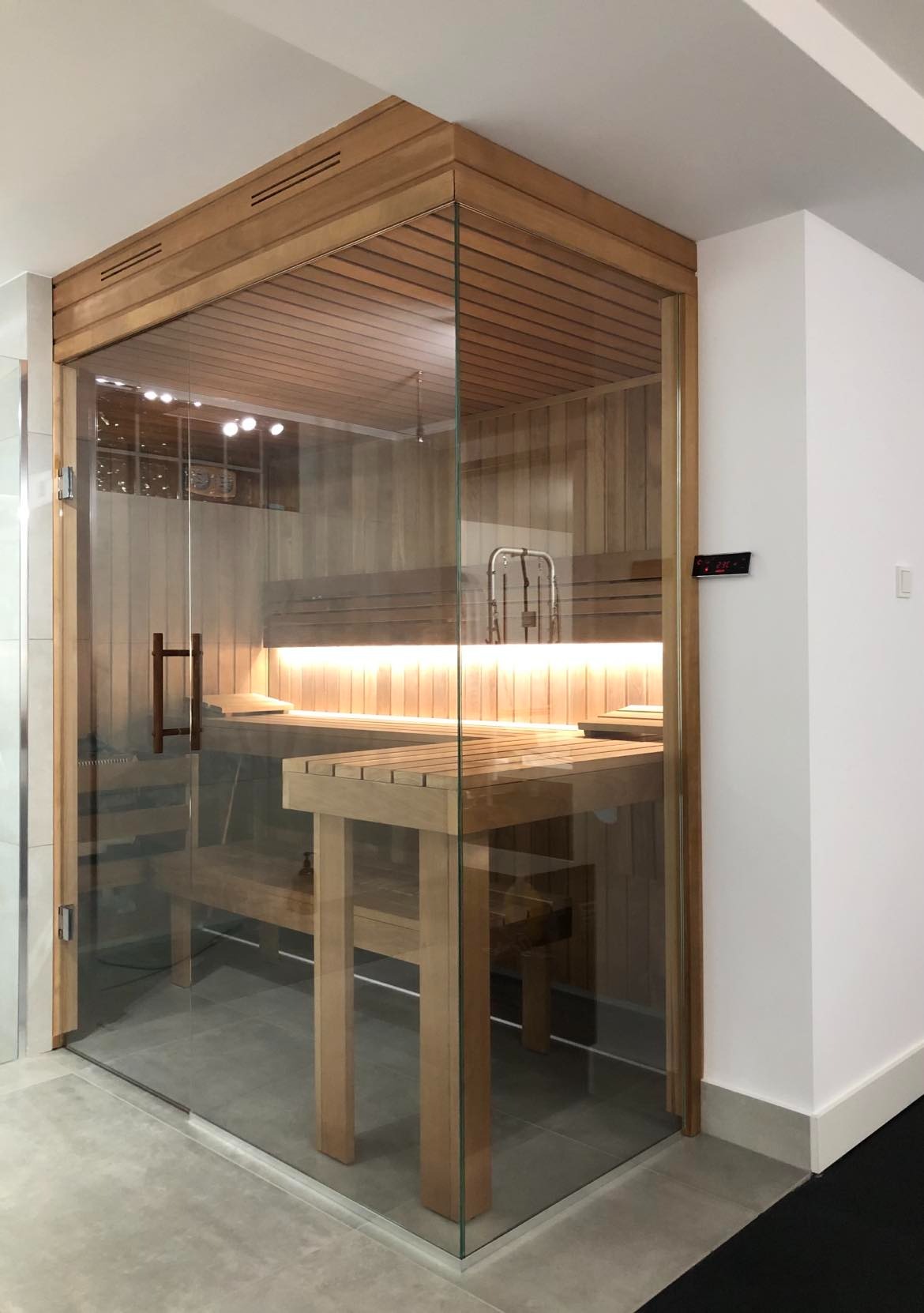 Nowoczesna, przeszklona kabina z drewnianą konstrukcją i ławkami, oświetlona listwą LED, z elektronicznym panelem sterowania na zewnątrz.