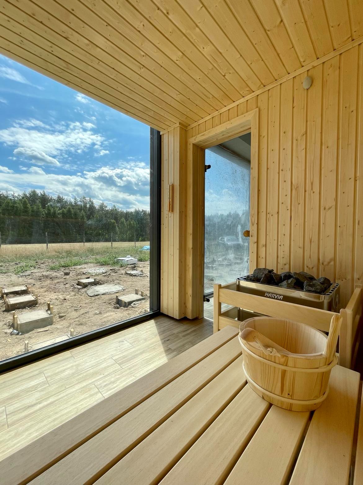 Wnętrze nowej sauny z jasnego drewna, widok na plac budowy przez panoramiczne okno, piec Harvia z kamieniami i drewniane wiadro z chochlą na ławce.
