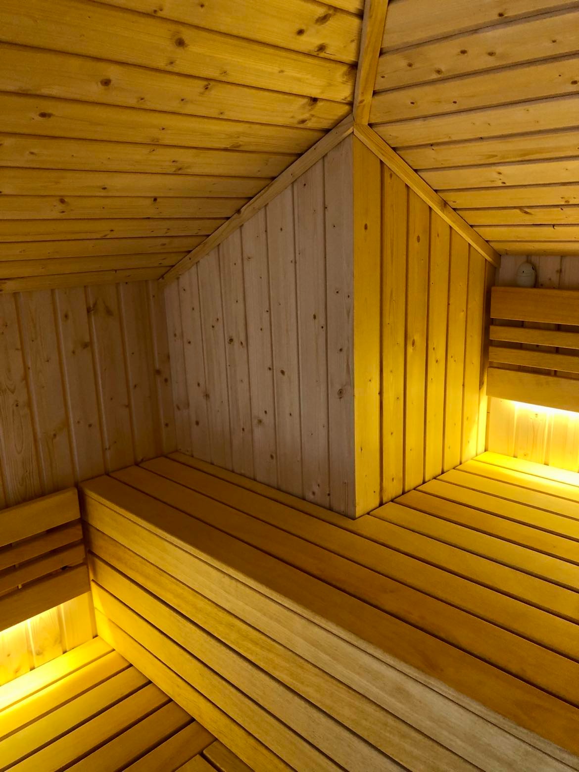 Wnętrze drewnianej kabiny z ławkami, oświetlone ciepłym światłem, widoczne połączenie jasnego i ciemnego drewna na ścianach i suficie.