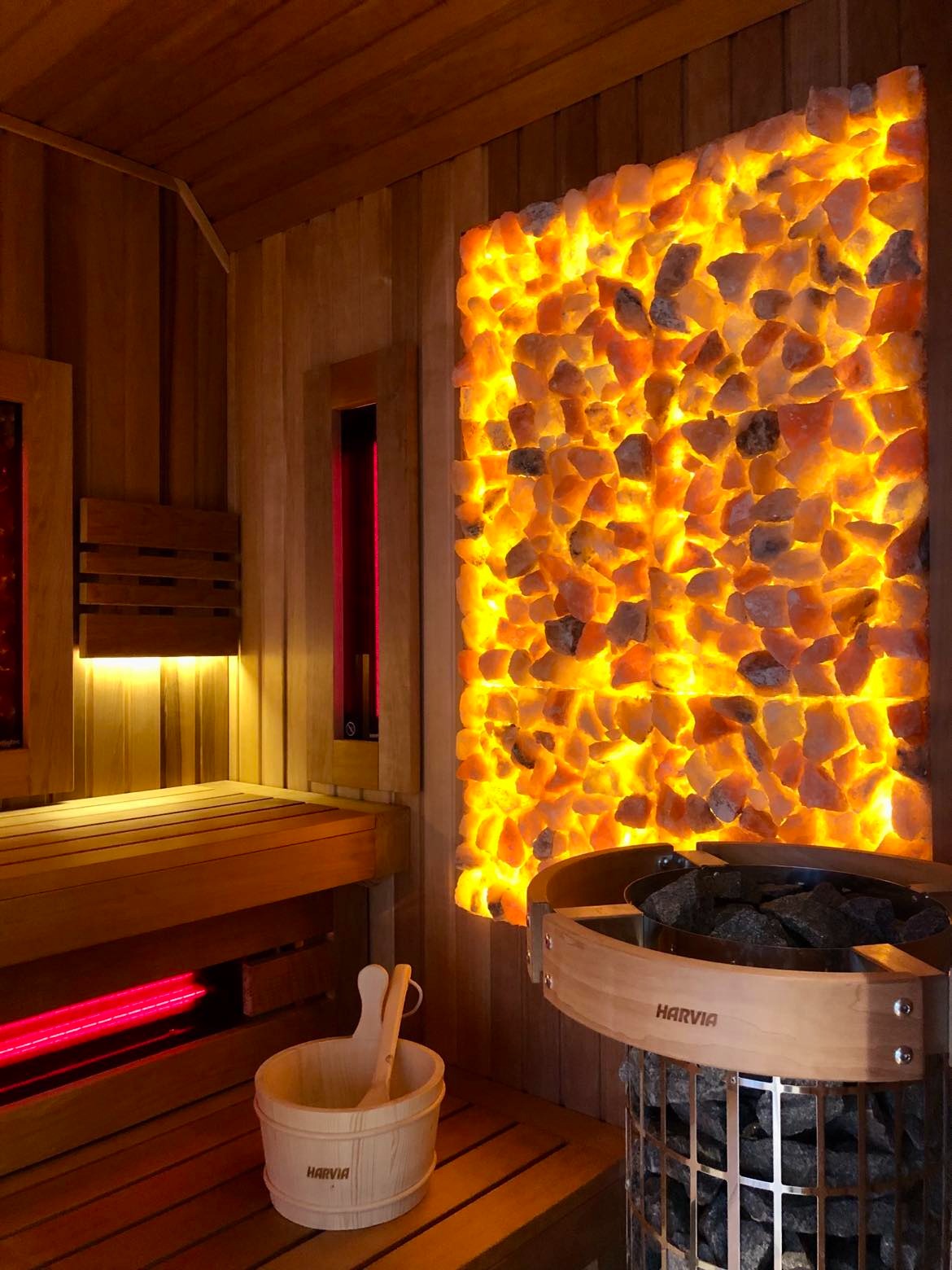 Drewniana sauna z podświetlaną ścianą z kamieni solnych, wiaderkiem i chochlą marki Harvia oraz piecem do sauny z kamieniami.