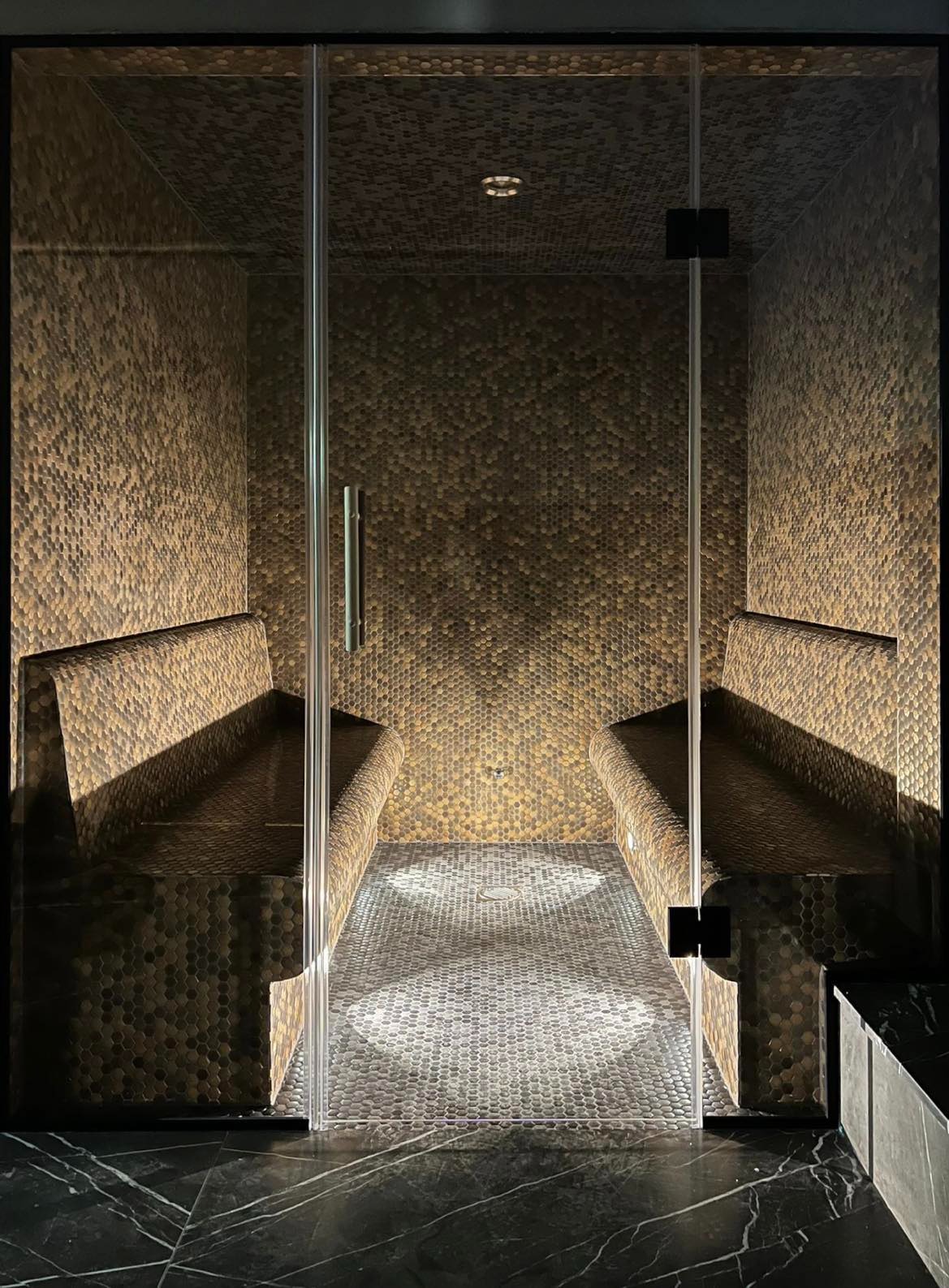 Wnętrze kabiny z mozaiką heksagonalną w odcieniach brązu i szarości, z dwoma ławami po bokach i szklanymi drzwiami, oświetlone punktowo