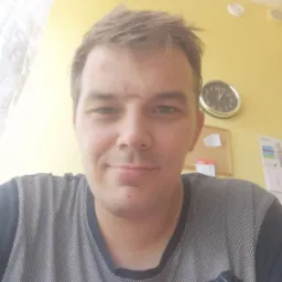 Portret mężczyzny w koszulce z geometrycznym wzorem na tle ż&oacute;łtej ściany z zegarem i kalendarzem.