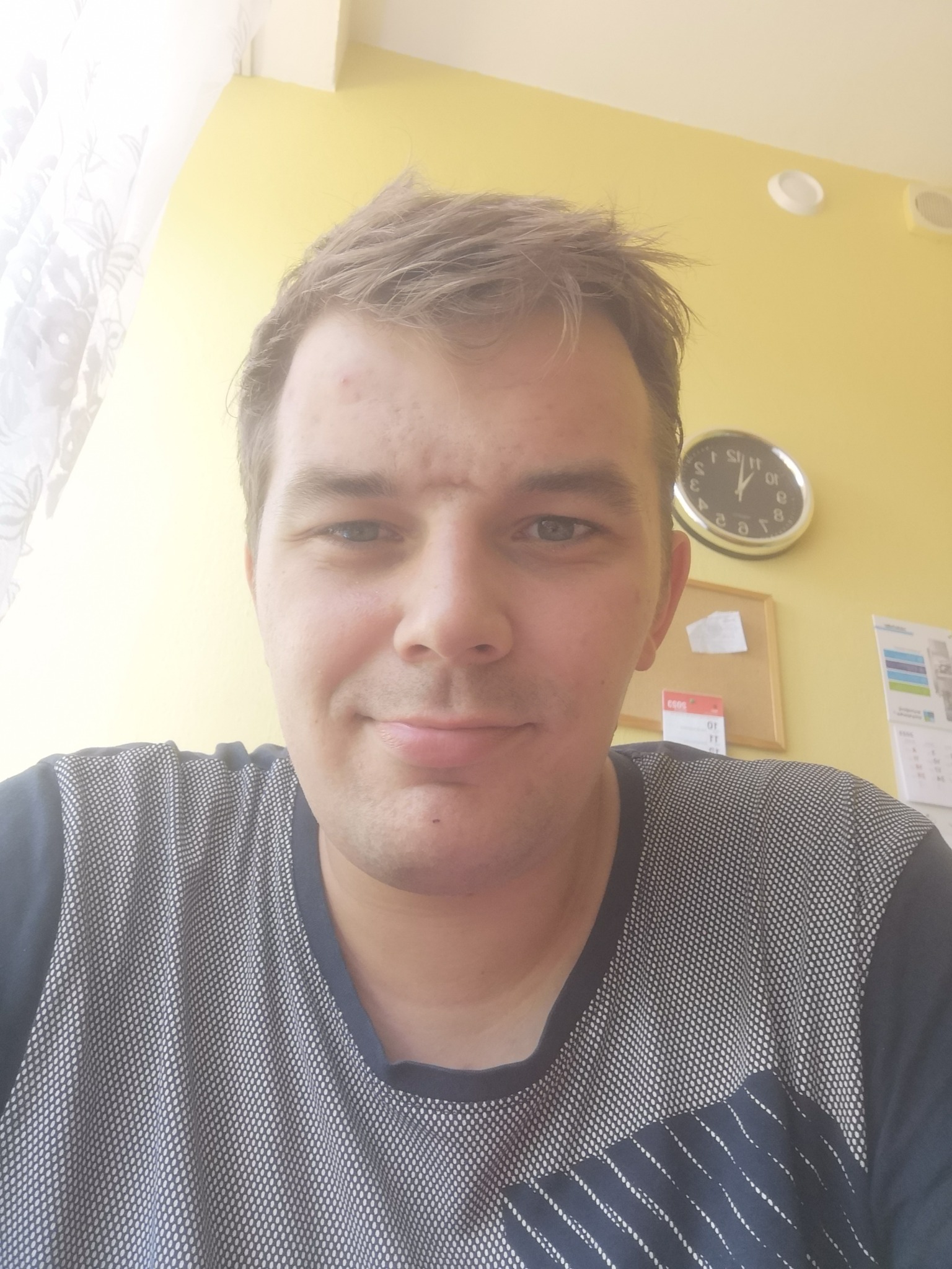 Portret mężczyzny w koszulce z geometrycznym wzorem na tle żółtej ściany z zegarem i kalendarzem.